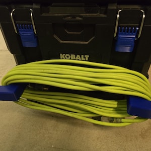 Op de afbeelding: Een zwarte Kobalt gereedschapskist met blauwe sluitingen en een opgerold, felgeel verlengsnoer. Het snoer wordt op zijn plaats gehouden door twee blauwe plastic eindstukken. De gereedschapskist staat op een betonnen vloer.