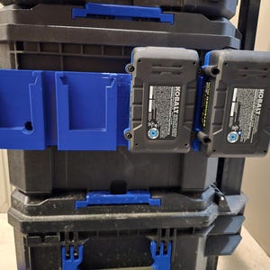 Puede incluir: Un soporte de batería azul impreso en 3D con dos ranuras, montado en una caja de herramientas negra. Dos baterías Kobalt grises y negras están adjuntas. La caja de herramientas tiene detalles en azul y está apilada con otras cajas de herramientas.