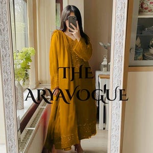 May include: Mustard yellow ensemble featuring a long-sleeved tunic and matching pants. The outfit is adorned with subtle embellishments and is reflected in a mirror. The text "THE ARYAVOGUE" is visible on the garment.