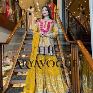 May include: A woman stands on a staircase wearing a yellow and pink embroidered outfit. The outfit includes a long-sleeved top with a pink neckline and a matching yellow skirt. The text "THE ARYAVOGUE" is visible.
