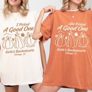 May include: Two t-shirts, one cream and one orange, with pumpkin graphics and text. The cream shirt reads "I Picked A Good One" and the orange shirt reads "She Picked A Good One." Both shirts say "Katie's Bachelorette, Chicago, IL, Est. 2025."