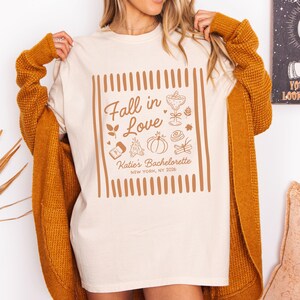 May include: A cream-colored t-shirt featuring a brown graphic print with the text Fall in Love, Katie's Bachelorette, and NEW YORK, NY 2026. The design includes illustrations of a cocktail glass, a pumpkin, a campfire, and cinnamon sticks.