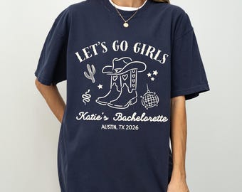 May include: Navy blue t-shirt with a white graphic that reads "LET'S GO GIRLS" with cowboy boots, a cactus, and a disco ball. The text "Katie's Bachelorette" and "AUSTIN, TX 2026" are also included.