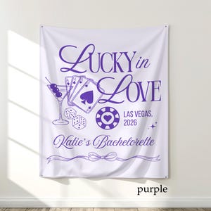 May include: White fabric banner with purple text and graphics. The banner reads "Lucky in Love" with Las Vegas, 2026, and "Katie's Bachelorette". Graphics include playing cards, dice, a martini, and a casino chip.