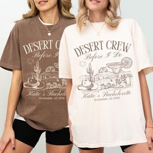 May include: Two t-shirts, one brown and one cream, with the text "DESERT CREW Before I Do" and "Katie's Bachelorette Scottsdale, AZ 2026". The graphic includes a desert scene with a cactus, hat, and margarita.