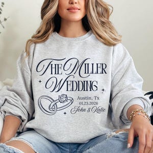May include: A light grey sweatshirt featuring the navy blue text "The Miller Wedding". The design includes two intertwined rings with a diamond, the location, date, and names: Austin, TX, 01.23.2026, John & Katie.