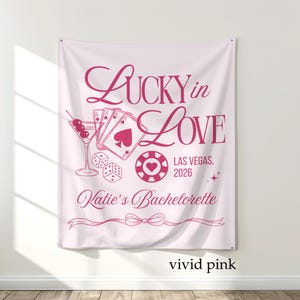 May include: Vivid pink tapestry with the text "Lucky in Love" and "Las Vegas, 2026". The design includes a martini glass, playing cards, dice, and a casino chip. The words "Katie's Bachelorette" are also included.