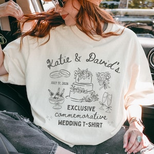 Op de afbeelding: Crèmekleurige sweatshirt met een gedenkwaardig huwelijksontwerp. Het ontwerp bevat de namen "Katie & David's", trouwringen, een taart, bloemen, champagne en de datum 17 juli 2026. De tekst "EXCLUSIVE commemorative WEDDING T-SHIRT" staat er ook op.