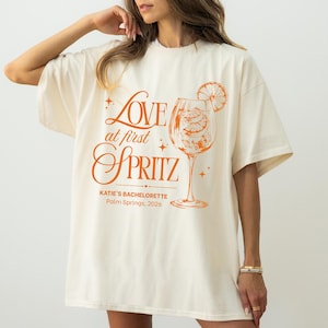 May include: Cream-colored oversized t-shirt with an orange graphic. The graphic features the words "Love at first Spritz" with a cocktail glass illustration and the text "Katie's Bachelorette, Palm Springs, 2026".
