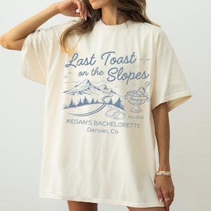 Könnte beinhalten: Cremefarbenes T-Shirt mit blauem Grafikdesign. Das Design enthält den Text "Last Toast on the Slopes", Berge, eine Straße, Champagner und den Text "Megan's Bachelorette, Denver, Co."