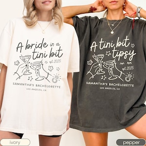 Op de afbeelding: Twee t-shirts, één ivoorkleurig en één peperkleurig, met de tekst "A bride in a tini bit" en "A tini bit tipsy". Beide shirts hebben een afbeelding van twee martiniglazen, bloemen, sterren en een ring, met "Samantha's Bachelorette" en "Los Angeles, CA".
