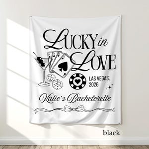 May include: White fabric banner with black text and graphics. The banner reads "Lucky in Love" with a martini glass, playing cards, dice, and a casino chip. The text "Las Vegas, 2026" and "Katie's Bachelorette" are also included.
