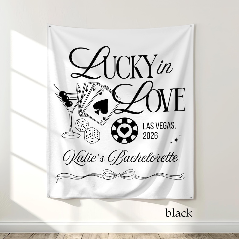 May include: White fabric banner with black text and graphics. The banner reads "Lucky in Love" with a martini glass, playing cards, dice, and a casino chip. The text "Las Vegas, 2026" and "Katie's Bachelorette" are also included.