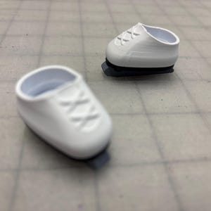 Puede incluir: Dos zapatillas blancas en miniatura con suelas grises se muestran sobre una superficie cuadriculada. Las zapatillas tienen un diseño clásico con cordones entrecruzados. Una zapatilla está enfocada, la otra está ligeramente borrosa.