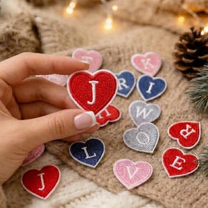 Puede incluir: Una mano sostiene un parche rojo en forma de corazón con la letra "J" bordada en blanco. Otros parches en forma de corazón con letras están dispersos sobre una superficie texturizada. Los parches son de varios colores, incluyendo rojo, rosa y azul.