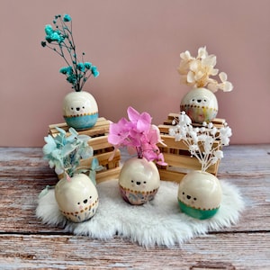 May include: Six small ceramic vases, each with a unique design and filled with dried flowers. The egg-shaped vases have painted faces and decorative bands of colour. The flowers are in shades of blue, pink, and white.