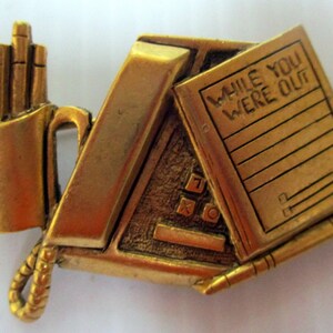 May include: Gold-toned metal brooch with a design of a pencil holder, a notepad with the text "While You Were Out", and a pen.