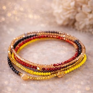 Puede incluir: Una colección de pulseras de cuentas en varios colores, incluyendo dorado, negro, rojo y amarillo. Las pulseras están dispuestas en un patrón circular, mostrando los diferentes colores y texturas de las cuentas. El fondo es un tono neutro suave y borroso.