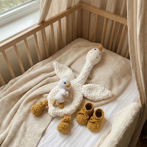 May include: A cream-colored crochet goose toy with a smaller duck toy, mustard yellow booties, and a cream-colored blanket. The toys are placed on a white sheet inside a wooden crib. The goose has a yellow beak and feet.