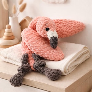 May include: A handmade pink flamingo plush toy with a white and gray beak, black eyes, and gray legs. The plush is made of a soft, textured material and is resting on a white blanket. The flamingo is a decorative item.