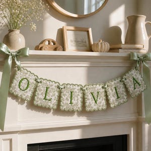 May include: A decorative fabric banner featuring the name OLIVIA in green embroidered letters on scalloped, cream-colored squares with a green floral pattern. The banner is suspended by light green satin ribbons tied into bows at each end.
