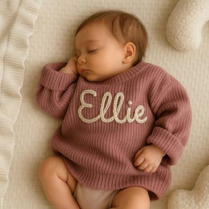 May include: A sleeping baby wearing a mauve-colored sweater with the name "Ellie" embroidered in white script across the chest. The sweater has ribbed cuffs and a crew neck. The baby is lying on a cream-colored surface.