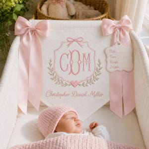 Könnte beinhalten: Ein weißes Stoffbanner mit rosa Stickereien, darunter ein Monogramm mit den Buchstaben CDM, ein Herz, florale Akzente und der Name Christopher Daniel Miller. Es ist mit zwei großen rosa Satinschleifen und einem Schild mit Zeilen für Date, Time, Weight, Length verziert.