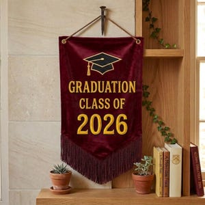 Puede incluir: Bandera de graduación de terciopelo granate con texto bordado en oro y flecos de borlas. La bandera dice "GRADUATION CLASS OF 2026" y presenta un birrete dorado. La bandera está colgada en una pared.