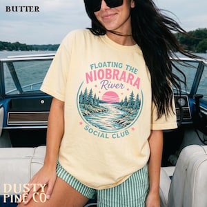 Puede incluir: Una camiseta de manga corta de color amarillo pálido con un gráfico circular que muestra un paisaje fluvial, pinos y un sol rosa. El texto dice Floating The Niobrara River Social Club. La camiseta se lleva con pantalones cortos a rayas verdes y blancas.
