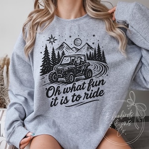 Może przedstawiać: Szara bluza z czarną grafiką UTV na krętej drodze, z drzewami, górami i słońcem. Tekst "Oh what fun it is to ride" znajduje się poniżej grafiki. Gwiazda i płatki śniegu również znajdują się w projekcie.