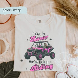 Puede incluir: Camiseta de color marfil con un gráfico de un vehículo todoterreno rosa y un conductor esqueleto. Los textos "Get in Loser" y "We're going Riding" están en escritura rosa. La camiseta es de material suave.