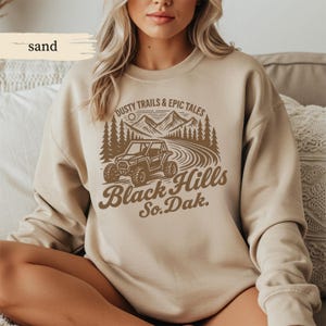 Może przedstawiać: Beżowa bluza z brązową grafiką. Grafika zawiera tekst "Dusty Trails & Epic Tales" i "Black Hills So. Dak.", scenę górską, drzewa i pojazd terenowy.