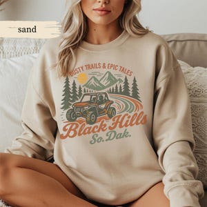 Op de afbeelding: Zandkleurige sweatshirt met een afbeelding van een rode terreinwagen, bergen, bomen en de tekst "Dusty Trails & Epic Tales Black Hills So. Dak." Het ontwerp heeft een vintage, outdoor esthetiek.