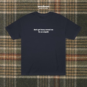 May include: Navy blue t-shirt with the text "don't get horny around me I'm an empath" in white. The brand name "wordloom" is printed at the top. The shirt is displayed on a plaid background.