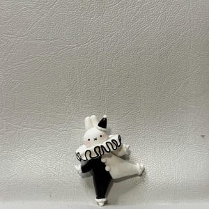 May include: A whimsical figurine of a rabbit dressed as a clown. The rabbit is primarily black and white, with a ruffled collar and a small hat. The figurine has a playful, cartoonish design.