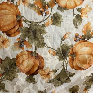 May include: A fabric with a repeating pattern of orange pumpkins, green leaves, and yellow flowers on a cream-colored background. The pumpkins have a textured appearance, and the overall design evokes a fall or harvest theme.