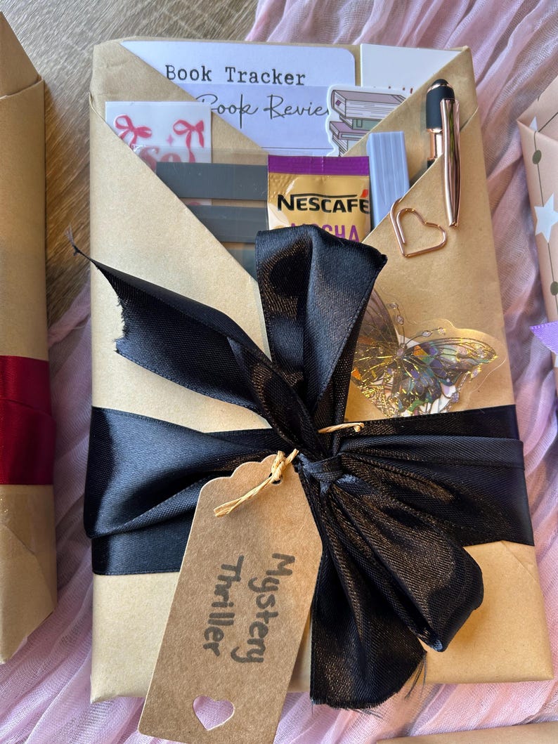 Op de afbeelding: Een bruin ingepakt cadeau, vastgebonden met een zwart lint en een label met de tekst "Mystery Thriller". Het bevat een pen, een hartvormige paperclip en een Nescafe-verpakking. Het zwarte lint is in een strik geknoopt.