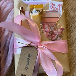 Op de afbeelding: Een cadeau verpakt in bruin papier en vastgebonden met een roze lint. Het bevat een "Book Tracker" kaart, een vlinderbedel en andere kleine items. Het label zegt "Romance".