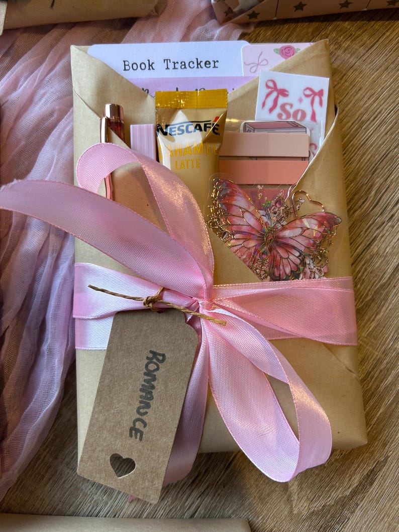 Op de afbeelding: Een cadeau verpakt in bruin papier en vastgebonden met een roze lint. Het bevat een "Book Tracker" kaart, een vlinderbedel en andere kleine items. Het label zegt "Romance".