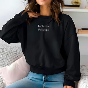 May include: Black sweatshirt with the text "For keeps? For keeps." in white. The sweatshirt has a crew neck and long sleeves. The model is wearing blue jeans.