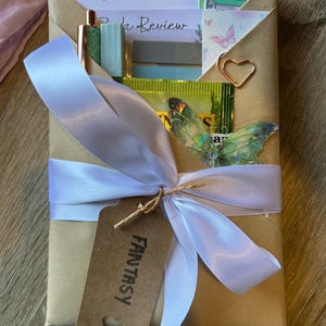 Op de afbeelding: Een cadeauverpakking met een wit lint en een bruin label met de tekst "FANTASY". De doos bevat briefpapier, een pen en een vlindersticker. De woorden "Book Tracker" en "Book Review" zijn zichtbaar.