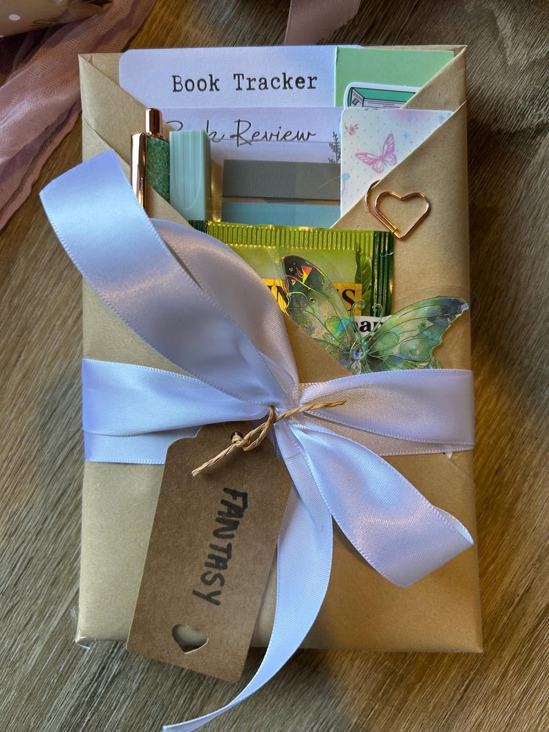Op de afbeelding: Een cadeauverpakking met een wit lint en een bruin label met de tekst "FANTASY". De doos bevat briefpapier, een pen en een vlindersticker. De woorden "Book Tracker" en "Book Review" zijn zichtbaar.