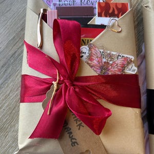 Op de afbeelding: Een cadeau verpakt in bruin papier, vastgebonden met een bordeauxrode lint en een label. Het pakket bevat een pen, stickers en een bladwijzer. Het label luidt "Dark Romance".
