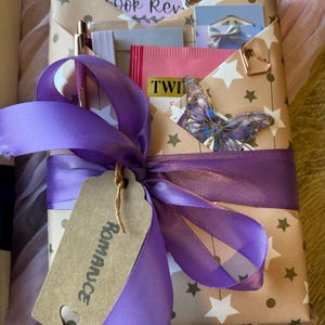 Op de afbeelding: Een cadeaupakket verpakt met een paars lint en een label met de tekst "Romance". Het bruine papier heeft witte sterren en bevat een boekenvolger, boekrecensiekaarten en een vlinderdecoratie.