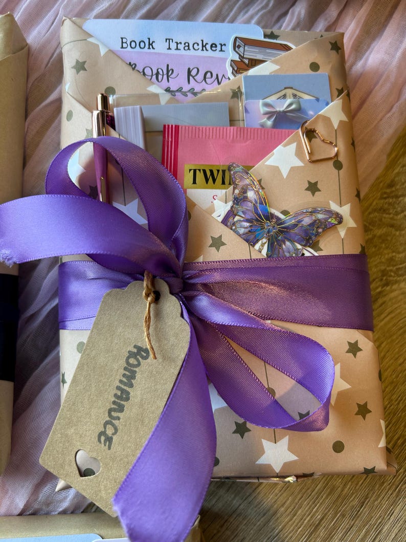Op de afbeelding: Een cadeaupakket verpakt met een paars lint en een label met de tekst "Romance". Het bruine papier heeft witte sterren en bevat een boekenvolger, boekrecensiekaarten en een vlinderdecoratie.
