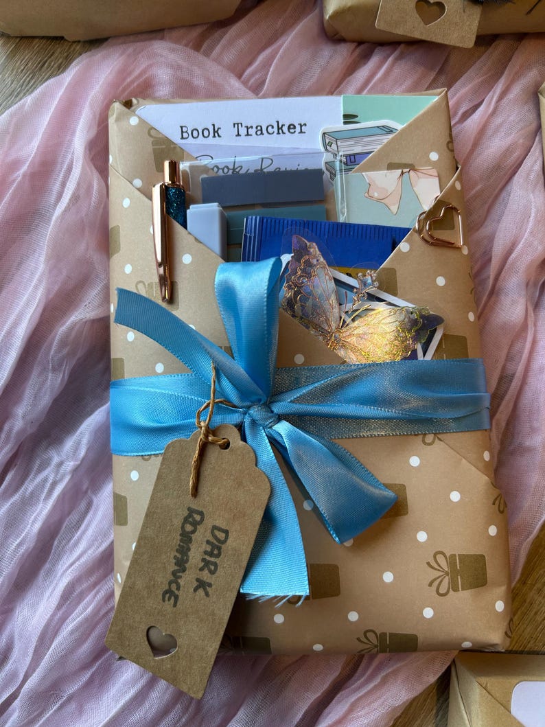 Op de afbeelding: Een ingepakt cadeau in bruin papier met witte stippen en gouden cadeau-ontwerpen, vastgebonden met een lichtblauw lint. Een label leest "Dark Romance". Een pen en andere items zijn zichtbaar. Het cadeau ligt op een roze ondergrond.