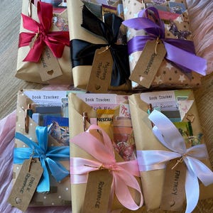 Op de afbeelding: Zes cadeaupakketten met boeken, verpakt in bruin papier, elk gebonden met een lint en label van een andere kleur. De linten zijn rood, zwart, paars, blauw, roze en wit. Labels lezen "Dark Romance", "Mystery Thriller", "Romance" en "Fantasy".