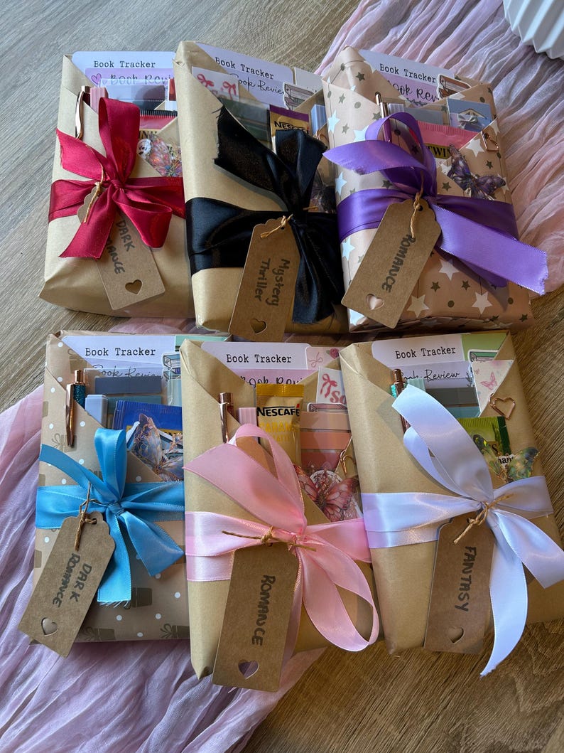 Op de afbeelding: Zes cadeaupakketten met boeken, verpakt in bruin papier, elk gebonden met een lint en label van een andere kleur. De linten zijn rood, zwart, paars, blauw, roze en wit. Labels lezen "Dark Romance", "Mystery Thriller", "Romance" en "Fantasy".