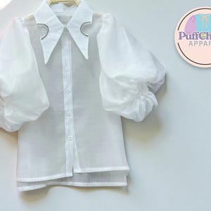 Könnte beinhalten: Weiße Bluse mit transparenten, puffärmeligen Ärmeln und einem Kragen. Der Kragen ist mit glitzernden Details verziert. Die Bluse hat eine Knopfleiste vorne und einen geschichteten Saum. Das PuffCharm Apparel Logo ist sichtbar.
