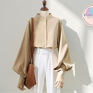 Può includere: Una camicetta beige con colletto alla coreana e maniche lunghe e voluminose è abbinata a pantaloni bianchi. Una borsa a tracolla marrone è appesa al manichino. Il logo PuffCharm Apparel è visibile in alto a destra.